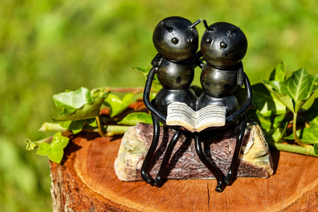 Statue of 2 ants sitting on a log with arms wrapped around shoulders as they read a book together.