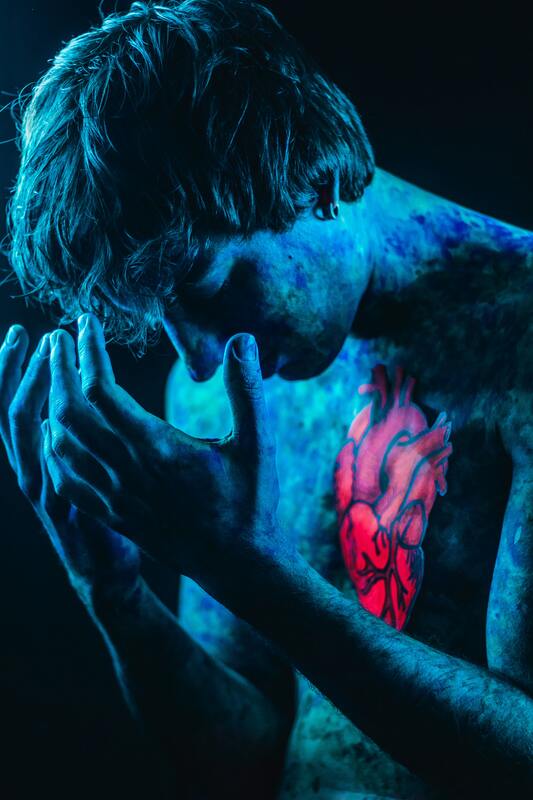 Human looking down, body painted in blue paint, a drawn red anatomical heart fixed to their chest