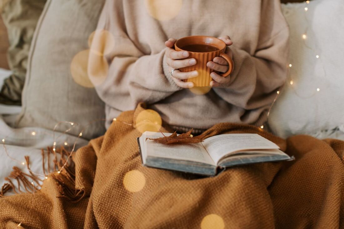 Person holding a warm drink with a blanket on their lap and an open book on the blanket