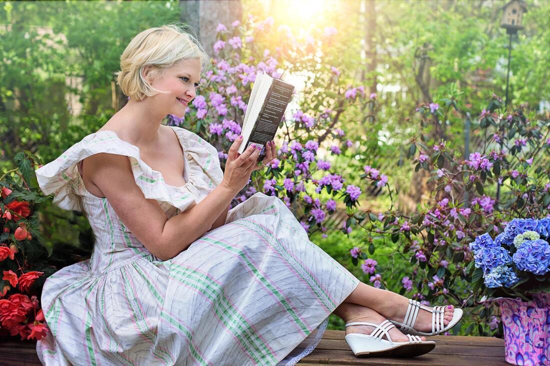 Woman wearing a spring dress joyfully reading a book while sitting in a flower garden